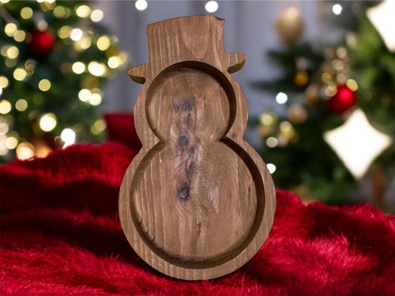 May include: A wooden snowman-shaped tray with a hat. The tray is made of natural wood and has a carved design. The tray is sitting on a red surface, with a Christmas tree in the background.