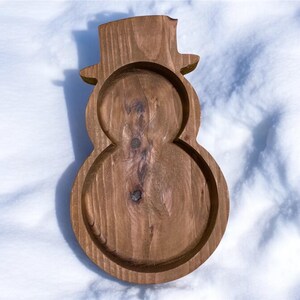 May include: A wooden snowman-shaped tray with a top hat design. The tray is made of light brown wood and has a carved-out design for holding items. The tray is sitting on a white snowy surface.