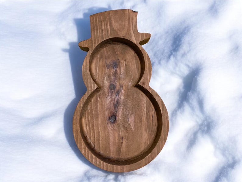 May include: A wooden snowman-shaped tray with a top hat design. The tray is made of light brown wood and has a carved-out design for holding items. The tray is sitting on a white snowy surface.