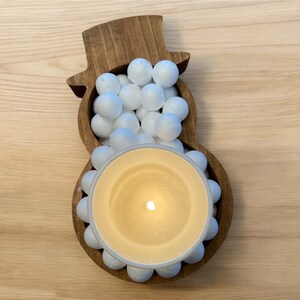 May include: A wooden snowman-shaped candle holder filled with white spheres and a lit candle. The candle is in a beige-colored container, and the snowman is made of brown wood.