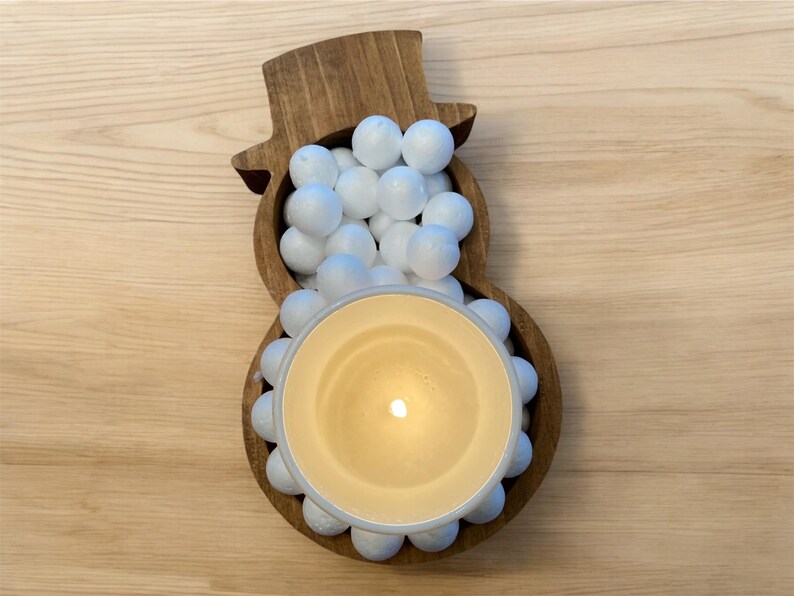 May include: A wooden snowman-shaped candle holder filled with white spheres and a lit candle. The candle is in a beige-colored container, and the snowman is made of brown wood.