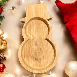 May include: Wooden snowman-shaped serving tray. The tray is made of light-colored wood and has a hat-shaped top. Christmas decorations, including ornaments and a red stocking, surround the tray.