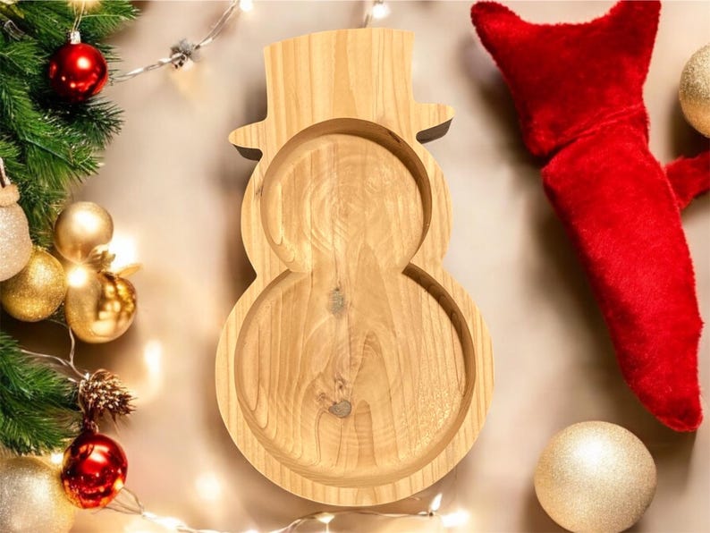 May include: Wooden snowman-shaped serving tray. The tray is made of light-colored wood and has a hat-shaped top. Christmas decorations, including ornaments and a red stocking, surround the tray.
