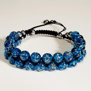 May include: A double-strand bracelet featuring blue glass beads with a speckled design. The beads are strung on a black cord with silver-colored metal accents and a sliding knot closure. The bracelet is displayed on a white surface.