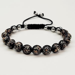 May include: Adjustable bracelet featuring black and gold speckled beads and silver-colored metal beads. The bracelet is strung on a black cord with a sliding knot closure. A stylish accessory for everyday wear.