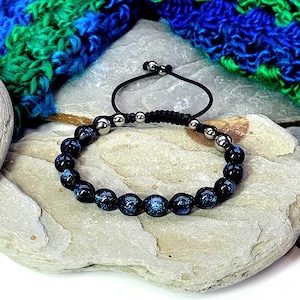 May include: A beaded bracelet featuring dark blue and silver beads. The bracelet is strung on a black cord with adjustable ends. The background includes a blue and green knitted item and a light-colored stone.