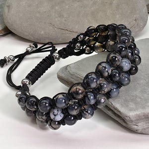 May include: A black beaded bracelet with gray and white accents. The bracelet is made of multiple strands of black cord, with silver-colored metal beads at the ends. The beads are a mix of dark gray, light gray, and white, creating a marbled effect.