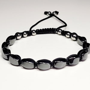 May include: A bracelet featuring a series of smooth, dark gray, rectangular beads strung on a black cord. The design includes smaller, round, metallic silver beads near the adjustable sliding knot closure at the back of the piece.