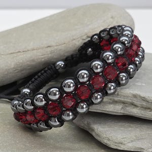 May include: A beaded bracelet featuring a combination of silver-colored beads and vibrant red crystals. The bracelet is crafted with a black woven cord and has an adjustable closure. The design is displayed on a gray stone surface.