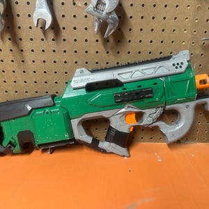 May include: Green and gray toy blaster with orange accents. The toy gun has a long barrel and a curved handle. The blaster is resting on an orange surface with tools hanging in the background.