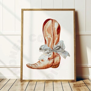 May include: Watercolor painting of a cowboy boot in shades of brown and red, adorned with a light blue bow. The artwork is framed in a wooden frame and displayed against a white background.