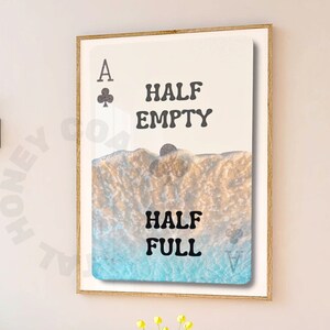May include: A framed art print featuring a playing card design with the Ace of Clubs symbol. The artwork displays the words "HALF EMPTY" and "HALF FULL" over a beach scene with blue and tan tones.