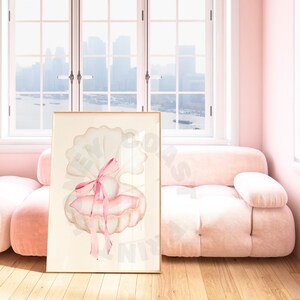 May include: A framed watercolor print featuring a seashell with a pearl and a pink bow. The artwork has a soft, pastel color palette of cream, pink, and beige. The print is displayed in a room with a pink sofa and a window.