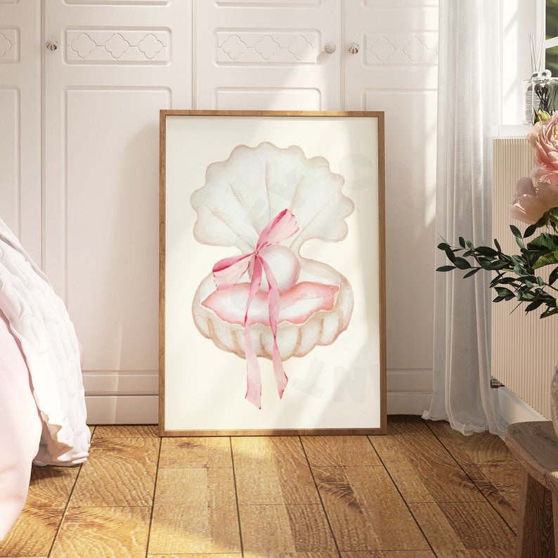 May include: Watercolor print of a seashell with a pearl and pink ribbon. The artwork is framed in a light brown wooden frame and is displayed on a wooden floor. The color palette is soft, with shades of white, pink, and beige.