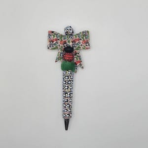May include: A decorative pen featuring a barrel covered in silver rhinestones and a top section adorned with a fabric bow patterned with Route 66 signs, cacti, and cars. The pen includes a green fuzzy bead, a red rhinestone bead, and a black bead.