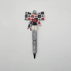 May include: A decorative pen featuring a black, white, and red checkered bow with small circular icons. The pen barrel is covered in silver rhinestones, accented by a white rhinestone bead, a black bead, a red rhinestone bead, and a silver topper.