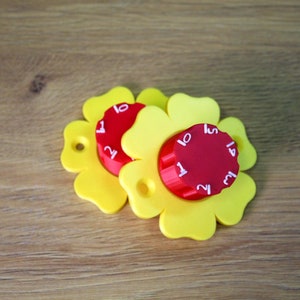 Könnte beinhalten: Zwei gelbe, blumenförmige Spielzeuge aus Plastik mit roten, runden Zifferblättern. Jedes Zifferblatt hat die Zahlen 1 bis 15 aufgedruckt.