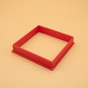 Può includere: Un tagliabiscotti quadrato rosso con base piatta e pareti verticali rialzate. L'oggetto presenta un semplice design geometrico con un centro cavo, costruito in materiale rosso solido con una visibile trama a strati sulle superfici.