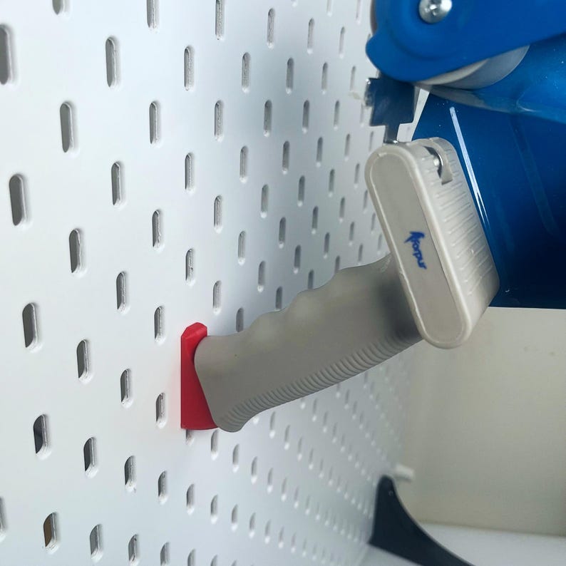 K&ouml;nnte beinhalten: Ein blauer und grauer Klebebandspender mit einem roten Haken, montiert an einer wei&szlig;en Lochwand. Der Spender hat einen grauen Griff und den Text "Mopur" auf der Seite.
