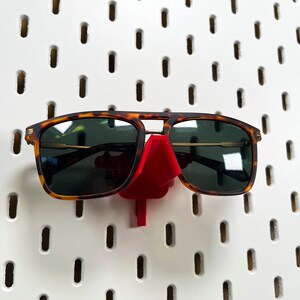 May include: A pair of tortoiseshell sunglasses with dark lenses are displayed on a red plastic sunglass holder. The holder is attached to a white pegboard.