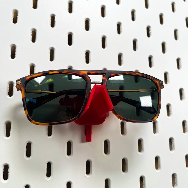 May include: A pair of tortoiseshell sunglasses with dark lenses are displayed on a red plastic sunglass holder. The holder is attached to a white pegboard.