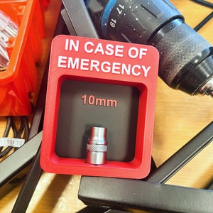 Könnte beinhalten: Eine rote Plastikbox mit dem Text "IM NOTFALL" und "10mm" darauf gedruckt. Eine silberne Steckschlüsselnuss befindet sich in der Box.