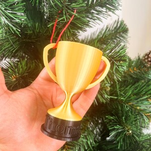 May include: A gold trophy ornament with a black base that says "Thomas" hanging on a green Christmas tree.