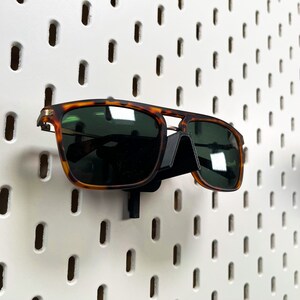 May include: Tortoise shell sunglasses with black frames and a black nose bridge. The sunglasses are hanging on a white pegboard.