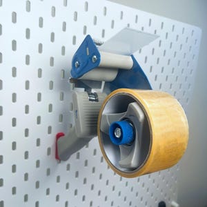 K&ouml;nnte beinhalten: Ein blauer und grauer Klebebandspender mit einer durchsichtigen Klebebandrolle. Der Spender ist an einer wei&szlig;en Lochwand montiert.