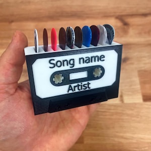 Könnte beinhalten: Ein weißer und schwarzer Kassettenband-förmiger Halter für Gitarrenplektren. Der Halter hat Schlitze für acht Plektronen und ein Etikett mit der Aufschrift "Song name" und "Artist".