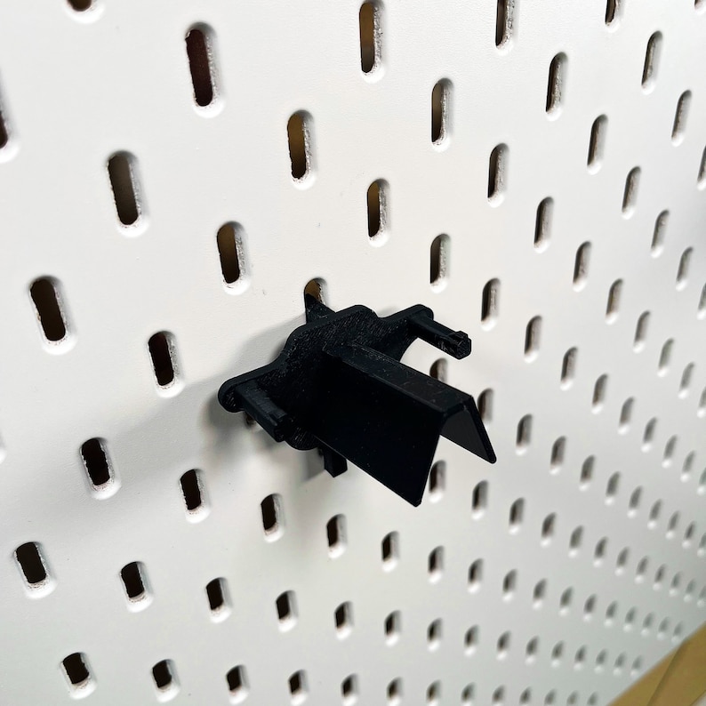 May include: Black plastic pegboard hook with a curved design. The hook is mounted on a white pegboard.