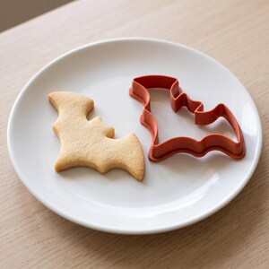 May include: A light brown bat-shaped cookie sits next to a matching orange plastic bat-shaped cookie cutter on a white plate. The cookie has a smooth surface and defined wing edges, while the cutter features a hollow center frame.