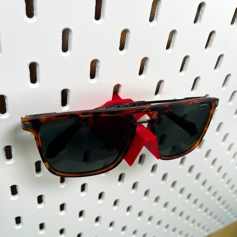 May include: Brown tortoiseshell sunglasses with a red plastic peg hook hanging on a white pegboard.