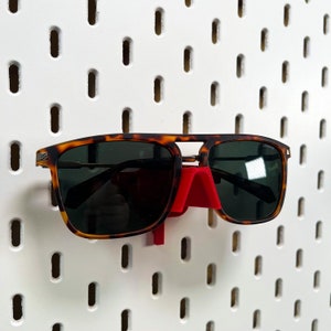 May include: A pair of tortoiseshell sunglasses with a red plastic hook hanging on a white pegboard.