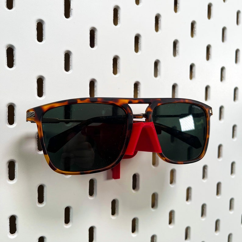 May include: A pair of tortoiseshell sunglasses with a red plastic hook hanging on a white pegboard.