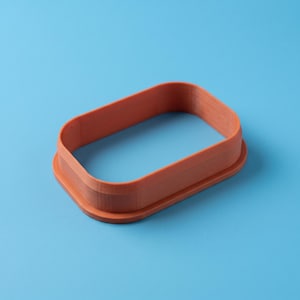 Può includere: Un tagliabiscotti rettangolare color terracotta con angoli arrotondati. L'oggetto presenta un bordo base largo e piatto e una struttura a parete verticale alta progettata per tagliare forme da impasti o altri materiali.