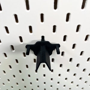 May include: Black plastic pegboard hook with a V-shaped bottom and two side arms. The hook is mounted on a white pegboard.