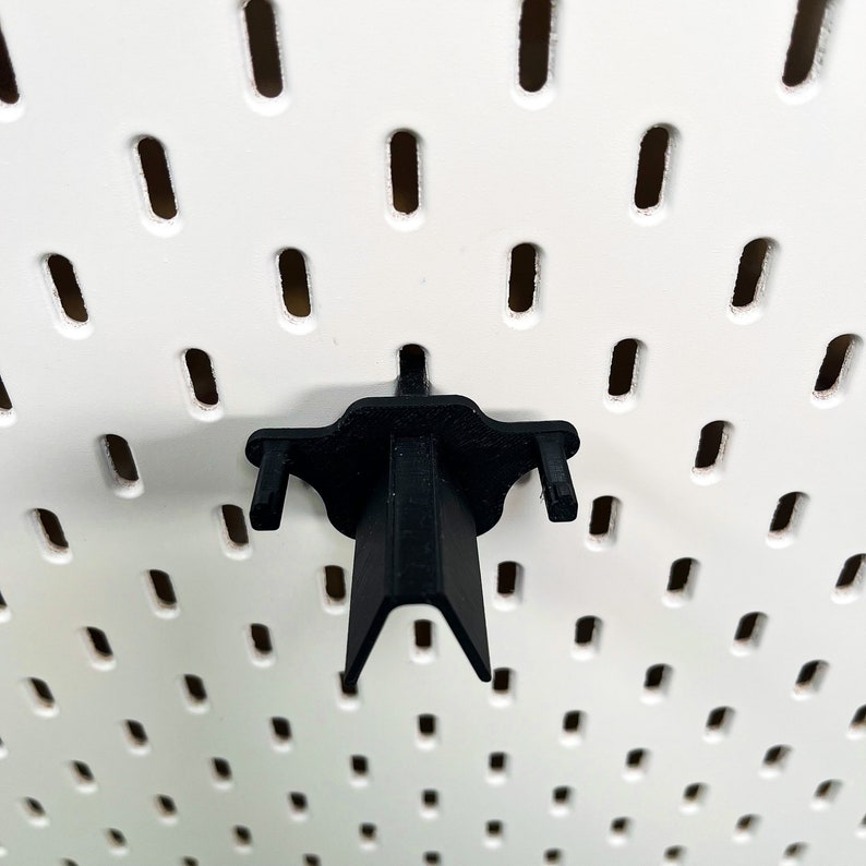 May include: Black plastic pegboard hook with a V-shaped bottom and two side arms. The hook is mounted on a white pegboard.