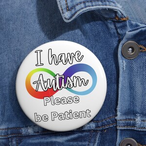 Op de afbeelding: Witte knop met de tekst "Ik heb autisme Wees geduldig" in het zwart. Het woord "Autisme" is omgeven door een regenboog oneindigheidssymbool.