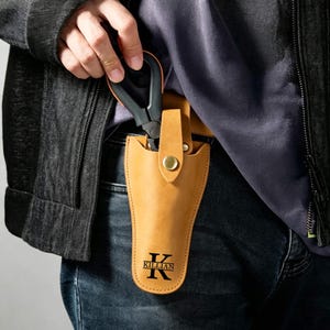 May include: A tan leather holster featuring a brass snap closure and a black printed logo with the letter K and the name KILLIAN. The holster is designed to hold a black and orange tool, which is partially inserted into the top opening.