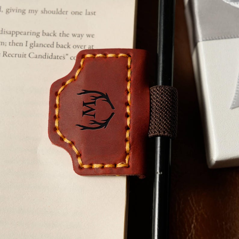 May include: A brown leather pen holder with a stitched border and a decorative antler design with the letter "M". The pen holder is attached to a black pen with a brown elastic band.