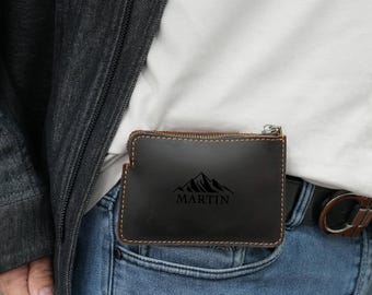 Puede incluir: Una cartera de cuero marrón con cierre de cremallera y un diseño de montaña con el nombre "MARTIN" grabado.