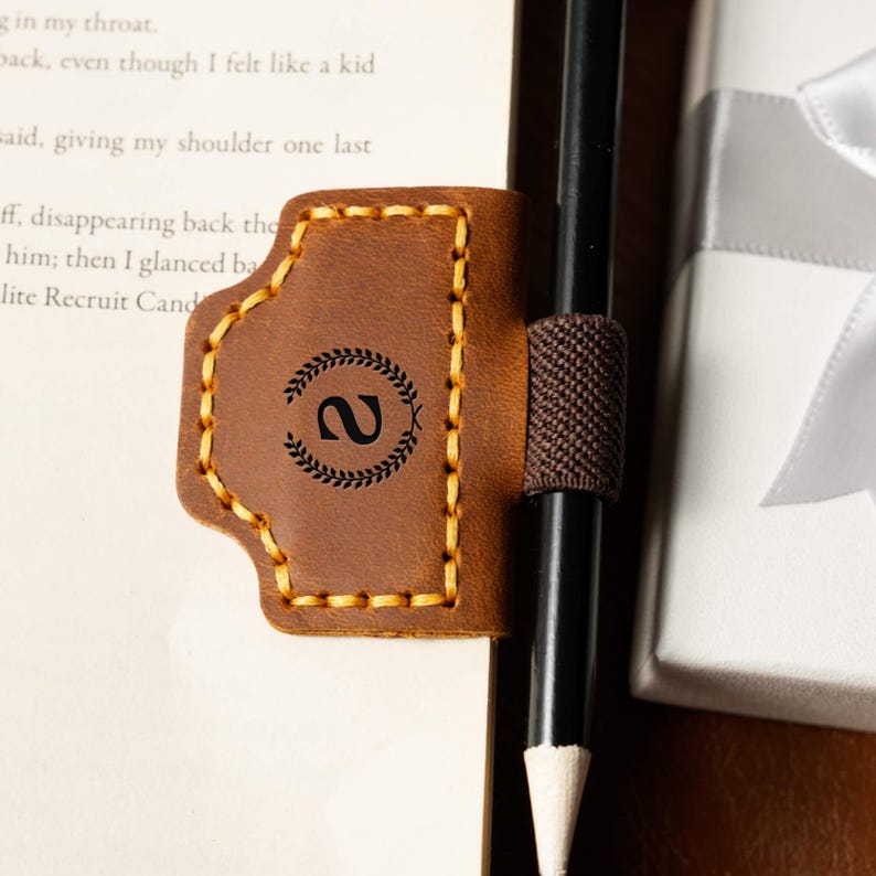 May include: A brown leather bookmark with a stitched border and a decorative wreath design with the number 2. The bookmark has a pencil holder and is next to a pencil and a book.