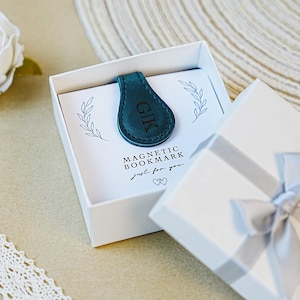 May include: A teal magnetic bookmark with the initials "GIK" in a white gift box. The box contains a card that reads "MAGNETIC BOOKMARK just for you" with leaf illustrations. The box is tied with a silver ribbon.