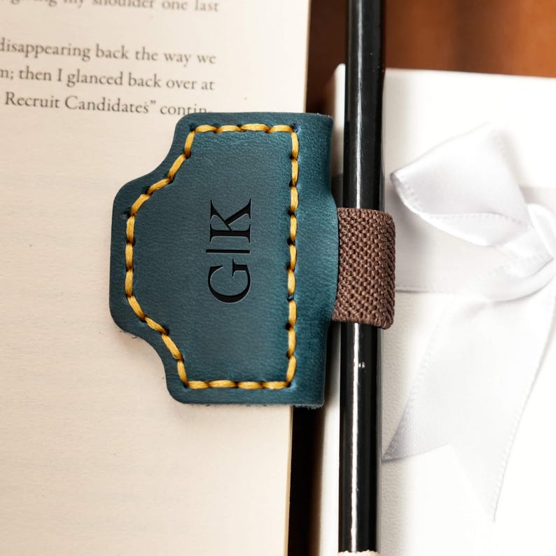 May include: Teal leather pen holder with yellow stitching and the initials "G|K" engraved. The pen holder is attached to a brown elastic band and holds a black pen. A white gift box is in the background.