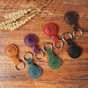 Pode incluir: Uma coleção de chaveiros de couro em várias cores, incluindo marrom, vermelho, verde e roxo. Cada chaveiro apresenta um design circular com um anel de metal e uma tira de couro. Alguns chaveiros têm iniciais ou símbolos.