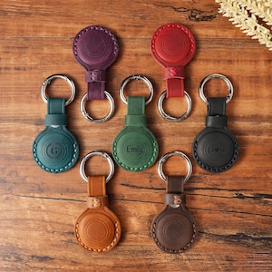 Puede incluir: Una colección de fundas de cuero para AirTag en varios colores, incluyendo verde, marrón y morado. Cada funda tiene un llavero plateado y está grabada con iniciales o un nombre. Las fundas están dispuestas sobre una superficie de madera.