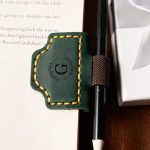 May include: A dark green leather pen holder with a gold stitched border and a brown elastic band. The holder features a debossed letter "G" within a laurel wreath design. A black pencil is held in place.