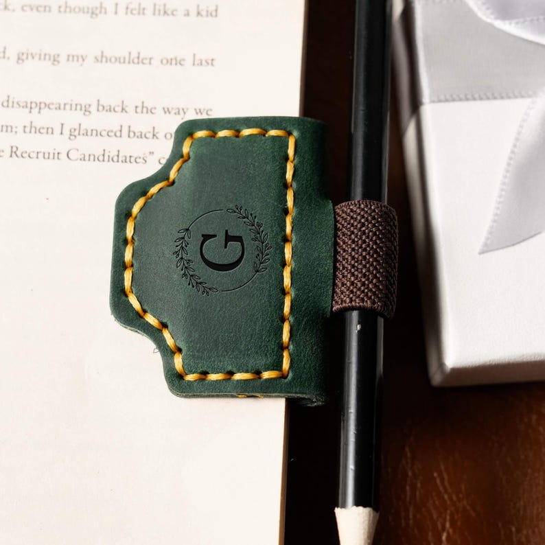 May include: A dark green leather pen holder with a gold stitched border and a brown elastic band. The holder features a debossed letter "G" within a laurel wreath design. A black pencil is held in place.