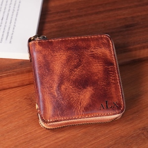 May include: A brown leather wallet with a zipper closure and the initials "ALX" embossed on the front. The wallet has a worn, textured appearance and a brass-colored zipper. The wallet is square and appears to be closed.